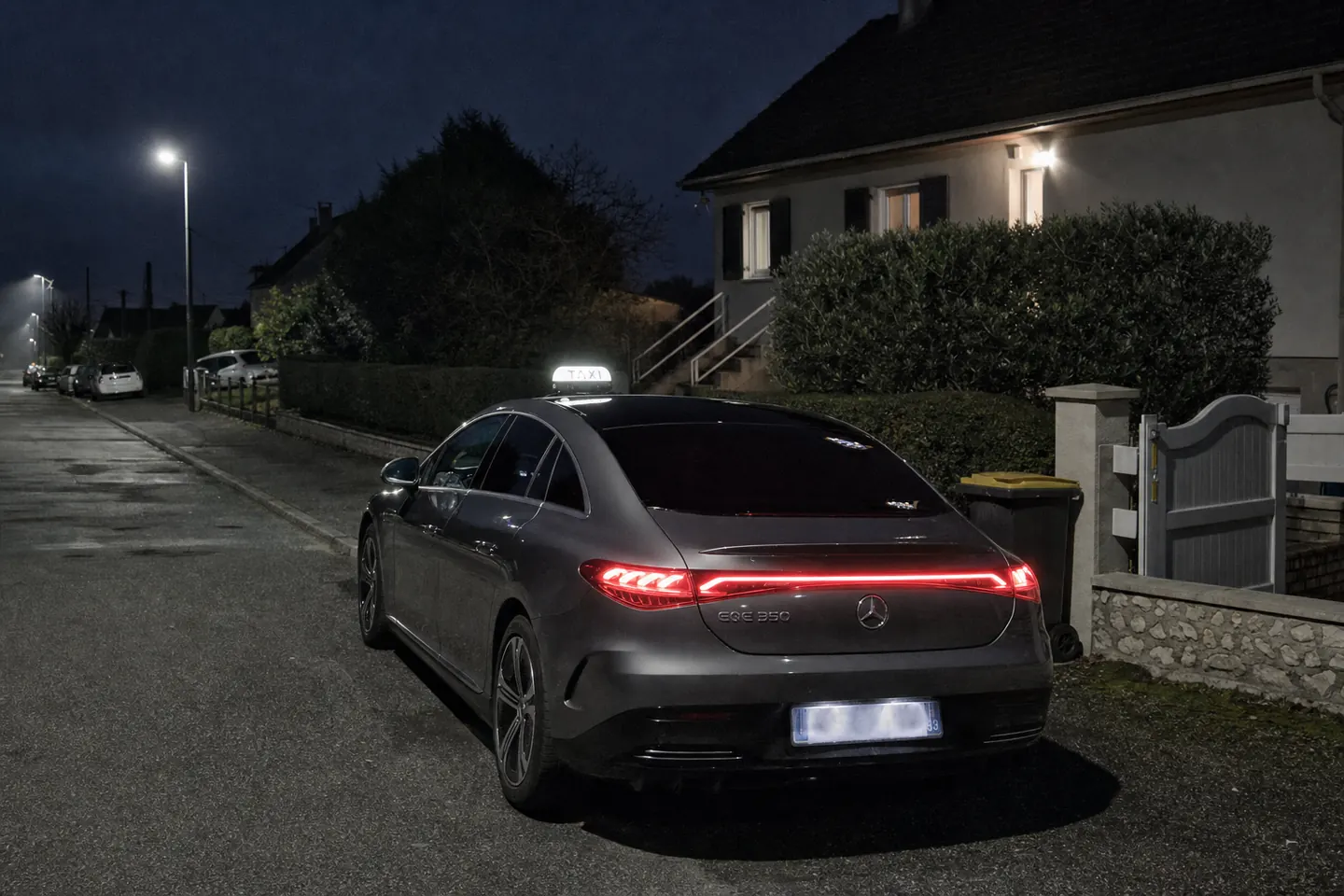 Taxi Mercedes EQE gris à l'arrêt devant un pavillon de la Goële en pleine nuit, lumineux allumé et feux arrière allumés