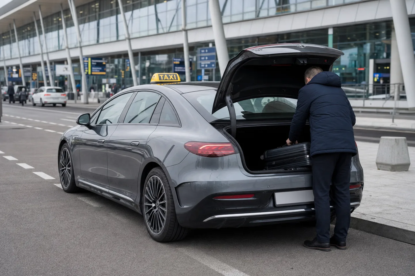 Taxi Mercedes EQE gris à l'arrêt le long du trottoir des arrivées d'un aéroport, coffre ouvert, un voyageur de dos y plaçant une valise
