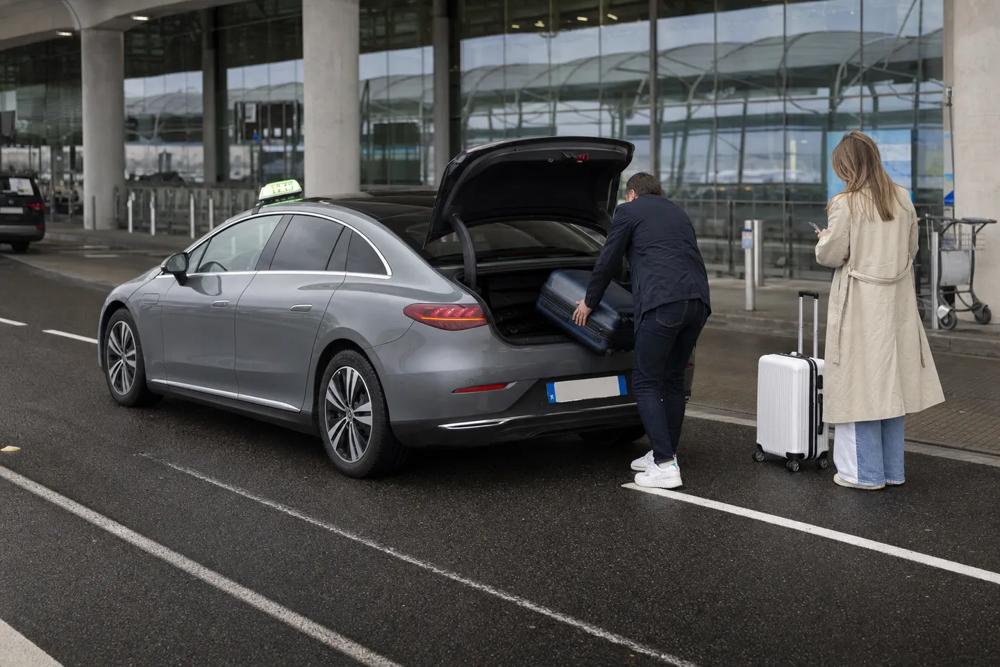 Taxi Mercedes EQE gris réservé à l'avance, coffre ouvert au trottoir des arrivées d'un terminal de Roissy, un voyageur y charge sa valise